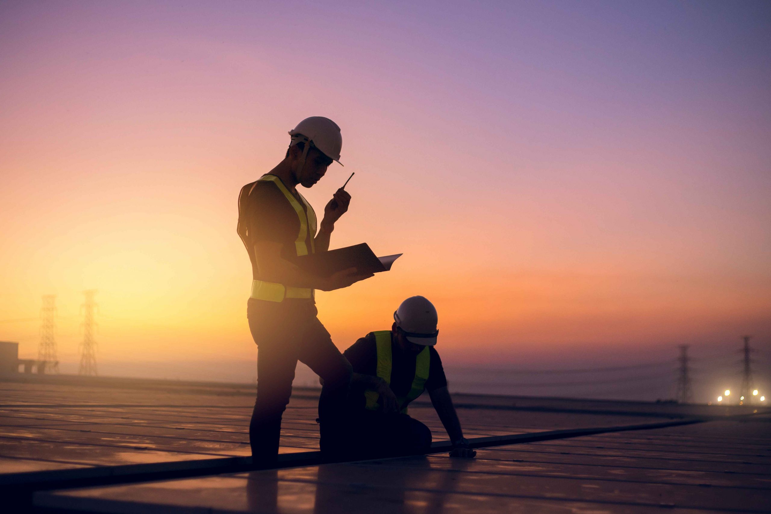 Two workers conducting a regular roof inspection at sunset, wearing safety vests and helmets, one reviewing notes while the other examines the roof