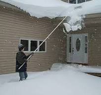 A person using a roof rake to remove heavy snowfall from the roof of a house, illustrating a common winter maintenance task
