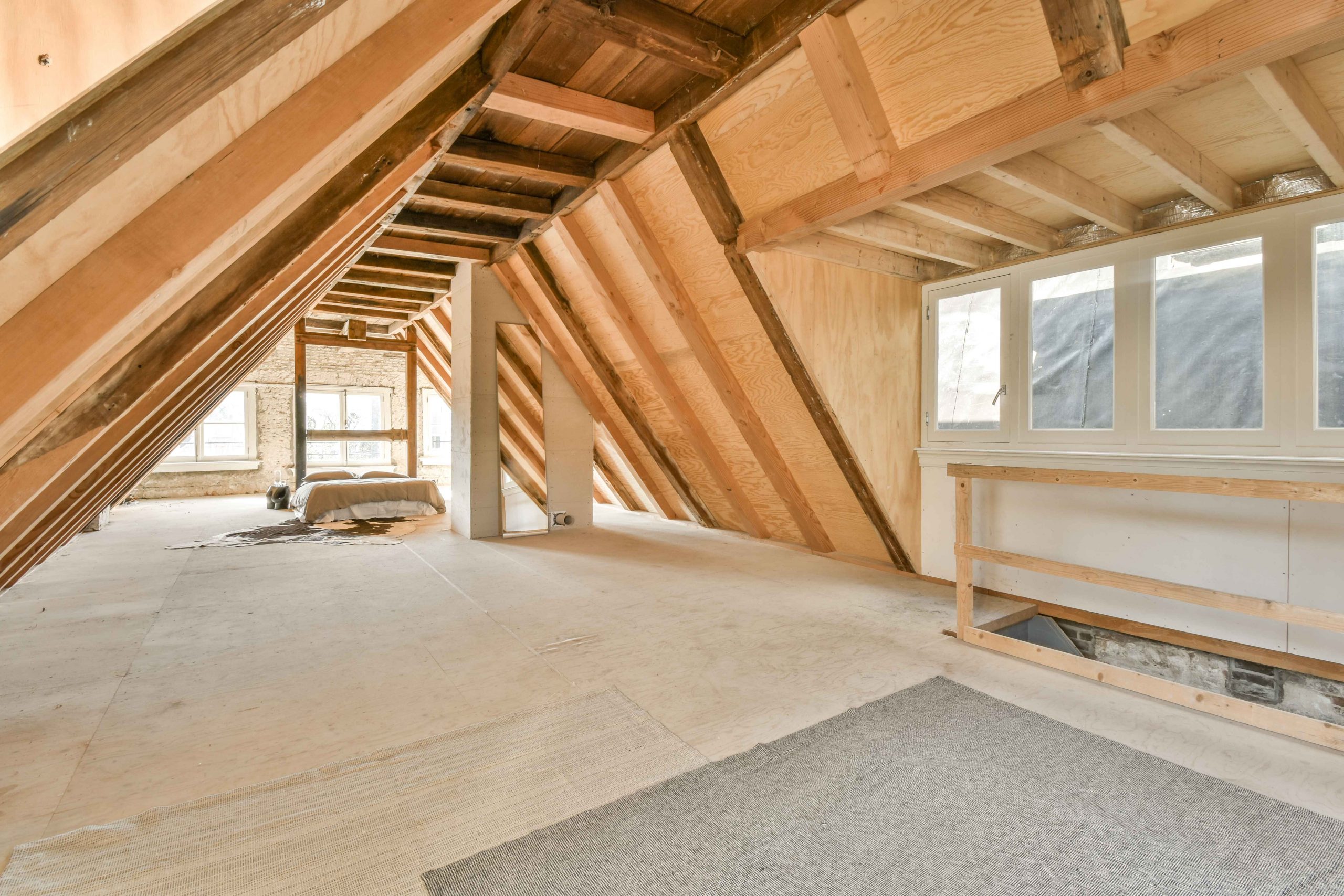 An unfinished attic space with exposed wooden beams and large windows, illustrating the importance of proper attic ventilation for maintaining a healthy roof structure