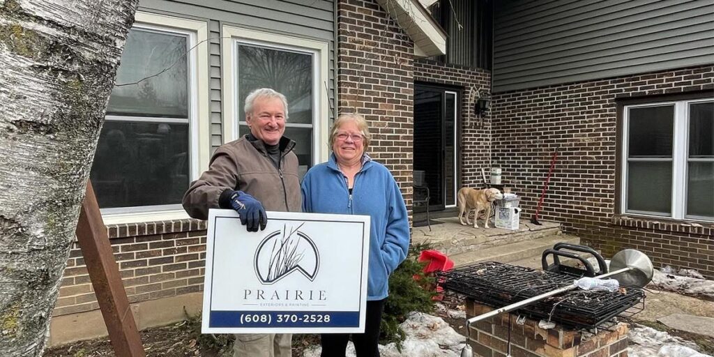Happy homeowners standing outside completed home exterior renovation project with Prairie Exteriors sign